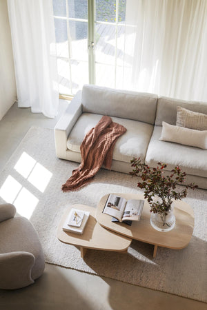 Ljust vardagsrum med beige soffa, ovalt soffbord i trä, terrakottafärgad filt och vita gardiner vid fönster