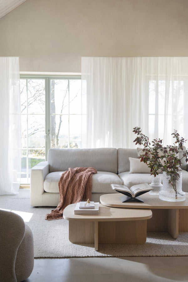 Ljust vardagsrum med beige soffa, runda soffbord i trä, vita gardiner och minimalistisk inredning