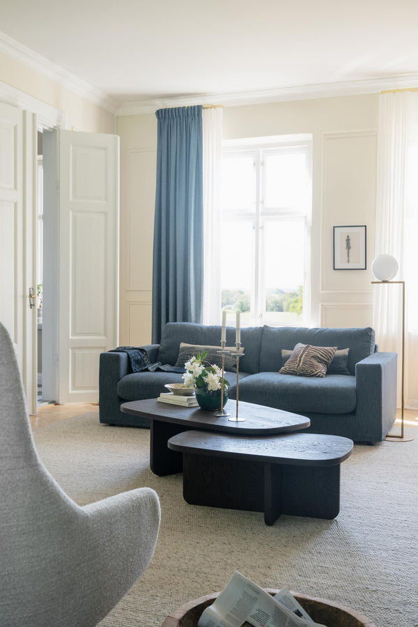 Modernt vardagsrum med blå soffa, svarta soffbord och ljusa väggar med fönster och gardiner