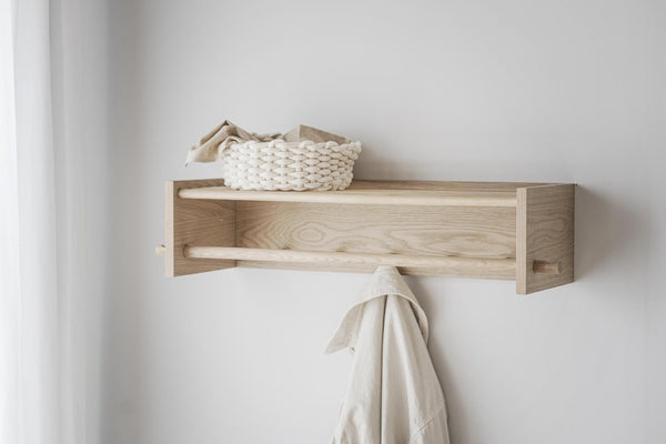 Ljus trähylla i ask med integrerad klädhängare monterad på vit vägg, dekorerad med flätad korg och handduk