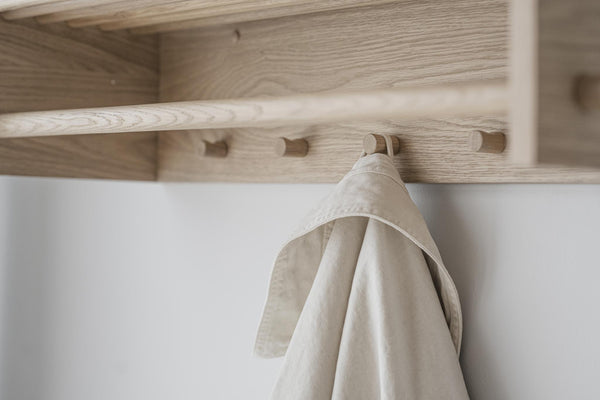 Klädhängare i ljust trä med beige linnekläder upphängda, monterad på vit vägg under hylla