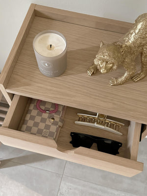 Sängbord i ljust trä med öppen låda innehållande solglasögon, klocka och plånbok, samt dekorativ guldpanterstatyett och do...