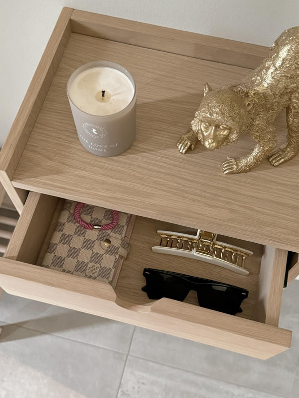 Sängbord i ljust trä med öppen låda innehållande solglasögon, klocka och plånbok, samt dekorativ guldpanterstatyett och do...