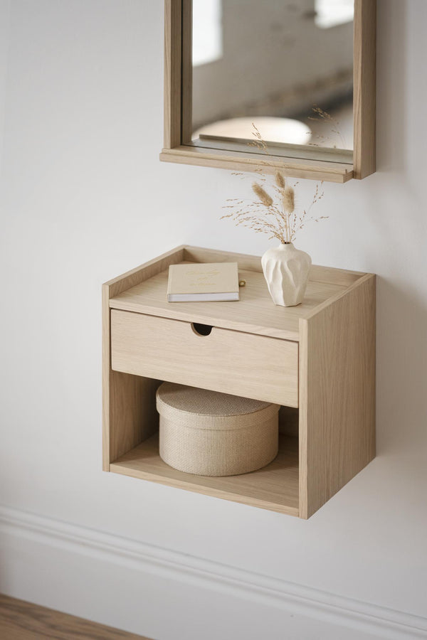 Väggmonterad sängbord i ljust trä med låda och öppen hylla, dekorerad med vas och torkade blommor