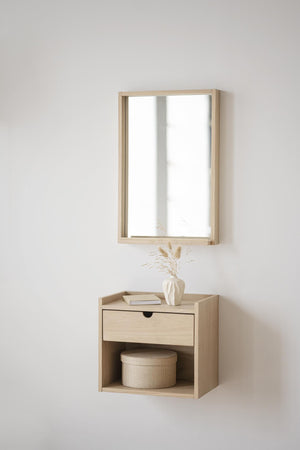 Väggmonterad spegel med träram ovanför sängbord i ljust trä med låda och öppen hylla