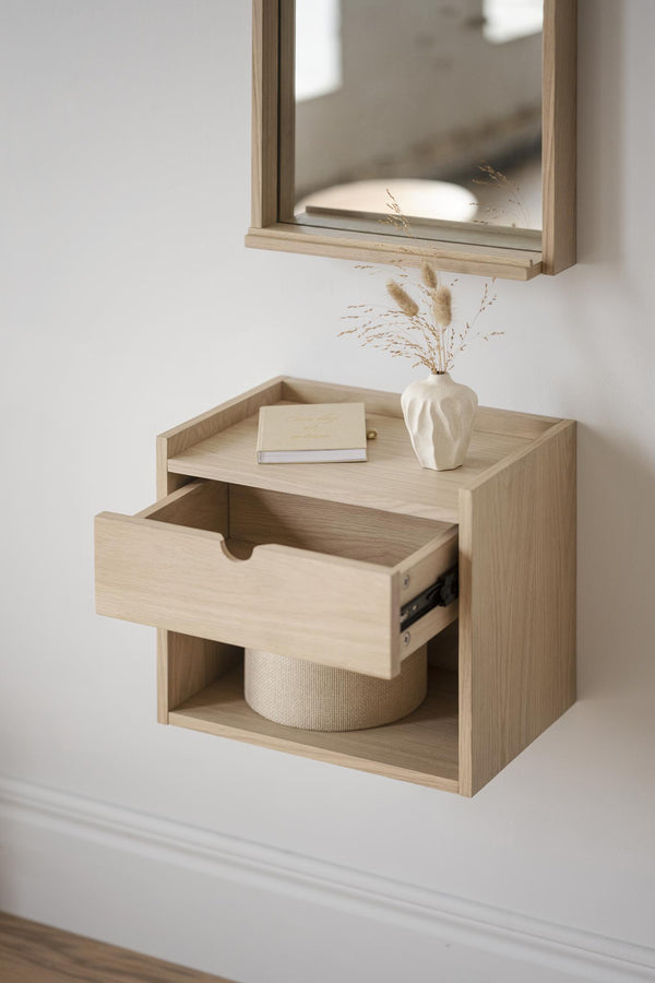 Väggmonterad sängbord i ljust trä med utdragen låda och öppen hylla, dekorerad med vas och bok
