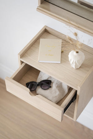 Sängbord i ljust trä med öppen låda innehållande solglasögon och anteckningsbok på hylla med dekorationer