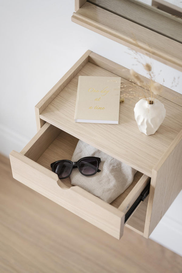 Sängbord i ljust trä med öppen låda innehållande solglasögon och anteckningsbok på hylla med dekorationer