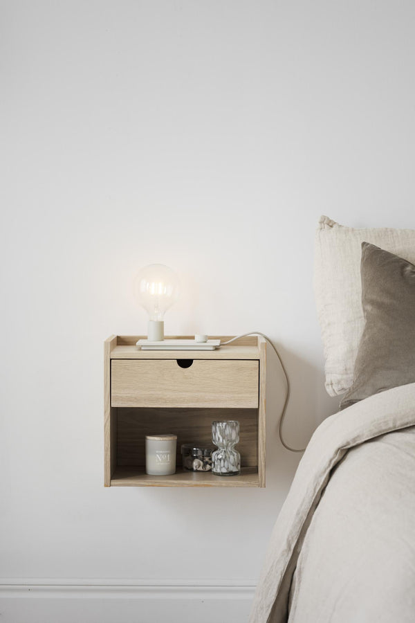 Väggmonterat sängbord i ljust trä med låda och öppen hylla, lampa och dekoration bredvid säng