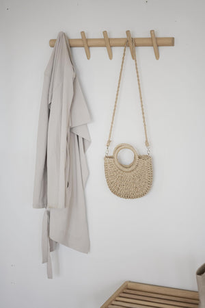Klädhängare i trä med fyra krokar på väggen, med grå kofta och en flätad väska i natur