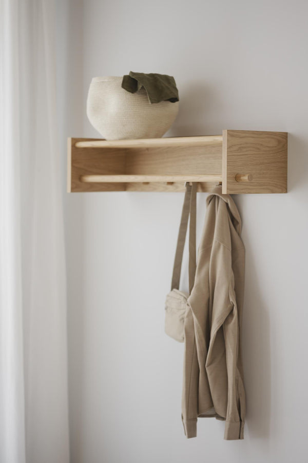 Väggmonterad hylla i ljust trä med krokfunktion, beige tygväska hängande och vit dekorativ skål