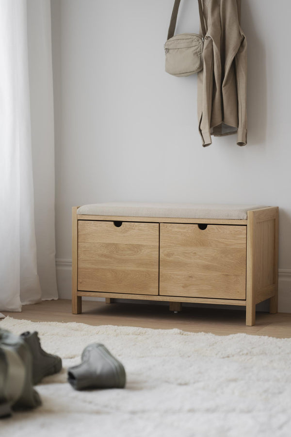 Hallbänk i ljust trä med stoppad sits och två förvaringslådor, placerad mot vit vägg med beige kläder hängande ovanför