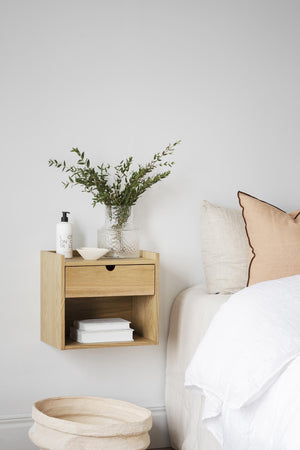 Väggmonterat sängbord i ek med låda och öppen hylla, dekorerat med vas och böcker vid vit säng