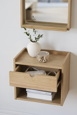 Väggmonterad sängbord i ek med utdragen låda, spegel ovanför, vit vas med gröna kvistar och glasskål på toppen