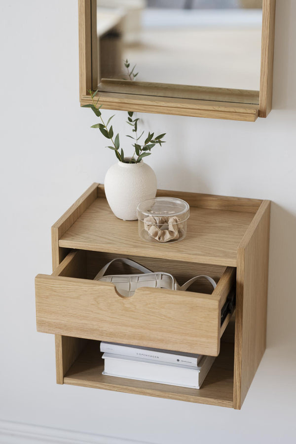 Väggmonterad sängbord i ek med utdragen låda, spegel ovanför, vit vas med gröna kvistar och glasskål på toppen