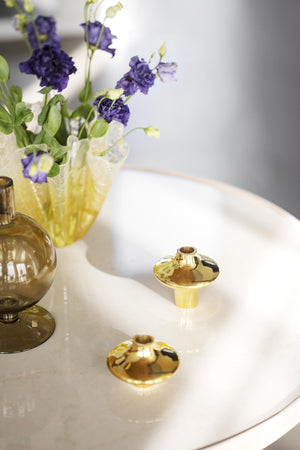 Guldiga ljusstakar i mässing på vit bricka tillsammans med lila blommor i glasvas