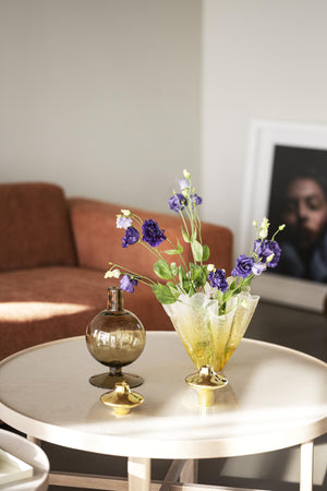 Glasvas med gula och lila blommor och rökfärgad glasflaska på vitt sidobord i vardagsrum