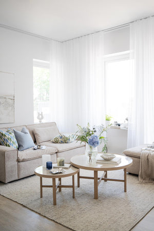 Ljust vardagsrum med beige soffa, runda soffbord i trä och vita genomskinliga gardiner vid fönster