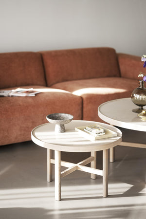 Runt soffbord i ljust trä med bricka, dekorativt fat och böcker framför brun soffa i vardagsrum