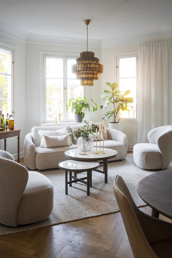 Ljust vardagsrum med beige boucléfåtöljer, runt soffbord, gyllene taklampa och gröna växter vid fönster