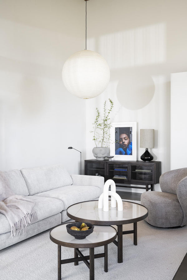 Modernt vardagsrum med vit taklampa, ljusgrå soffa, runda soffbord och svart skänk med tavla