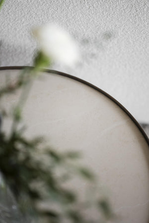 Rund spegel med svart ram och suddig grön växt i förgrunden mot vit vägg