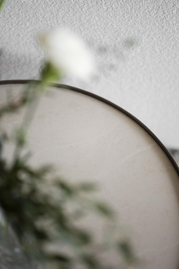 Rund spegel med svart ram och suddig grön växt i förgrunden mot vit vägg