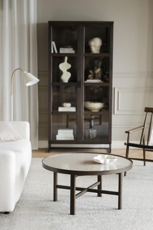 Runt soffbord i mörkt trä med vit skål, omgivet av vit soffa, golvlampa och vitrinskåp i vardagsrum