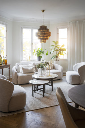 Ljust vardagsrum med beige boucléfåtöljer, rund soffbord och gyllene taklampa i takvåning