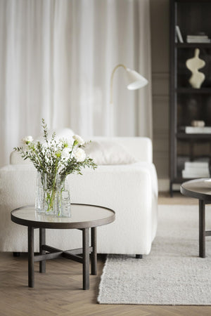 Runt soffbord i mörkt trä med glasskiva, vit boucléfåtölj och vas med vita blommor i modern skandinavisk inredning