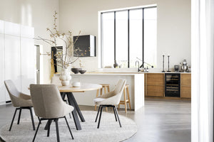 Ljust kök med matplats, runt matbord, beiga stolar och träskåp med vita bänkskivor