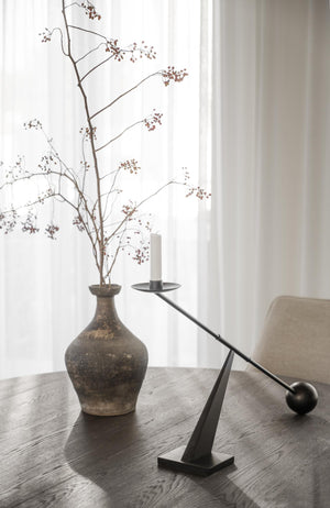 Minimalistisk ljusstake i svart metall med balansdesign bredvid antik vas med torkade grenar på träbord