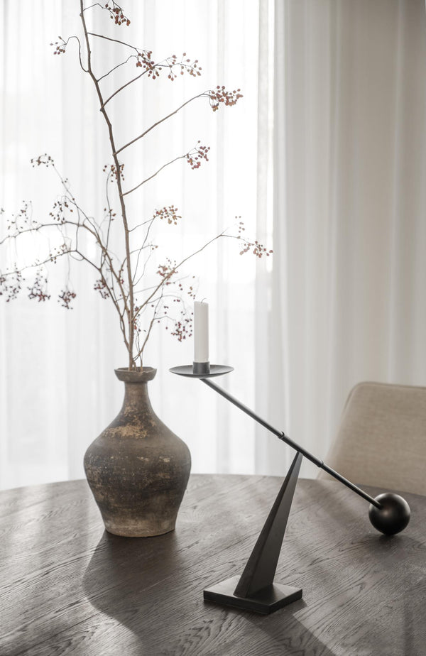 Minimalistisk ljusstake i svart metall med balansdesign bredvid antik vas med torkade grenar på träbord