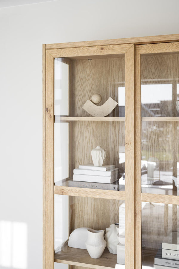 Vitrinskåp i ek med glashyllor och dekorativa vita keramikobjekt, minimalistisk skandinavisk design