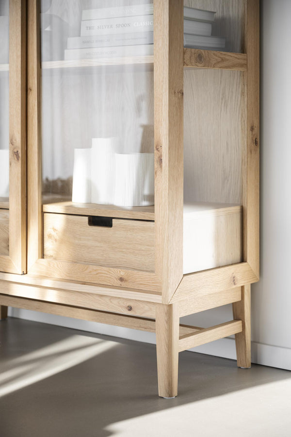 Vitrinskåp i ljust trä med glasdörrar, drawer och synliga föremål på hyllor