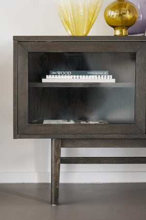 Sängbord i mörkgrått trä med glasdörr och synlig hylla, dekorerat med gula glasföremål