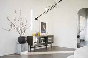 Minimalistisk inredning med skänk i mörkt trä, taklampa och dekorativa grenar i svart vas på betongpelare