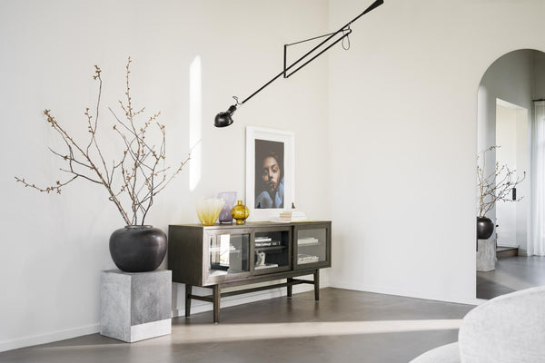 Minimalistisk inredning med skänk i mörkt trä, taklampa och dekorativa grenar i svart vas på betongpelare