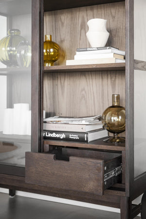 Bokhylla i mörkt trä med glasdörr, öppna hyllplan, utdragen låda och dekorativa böcker och vaser