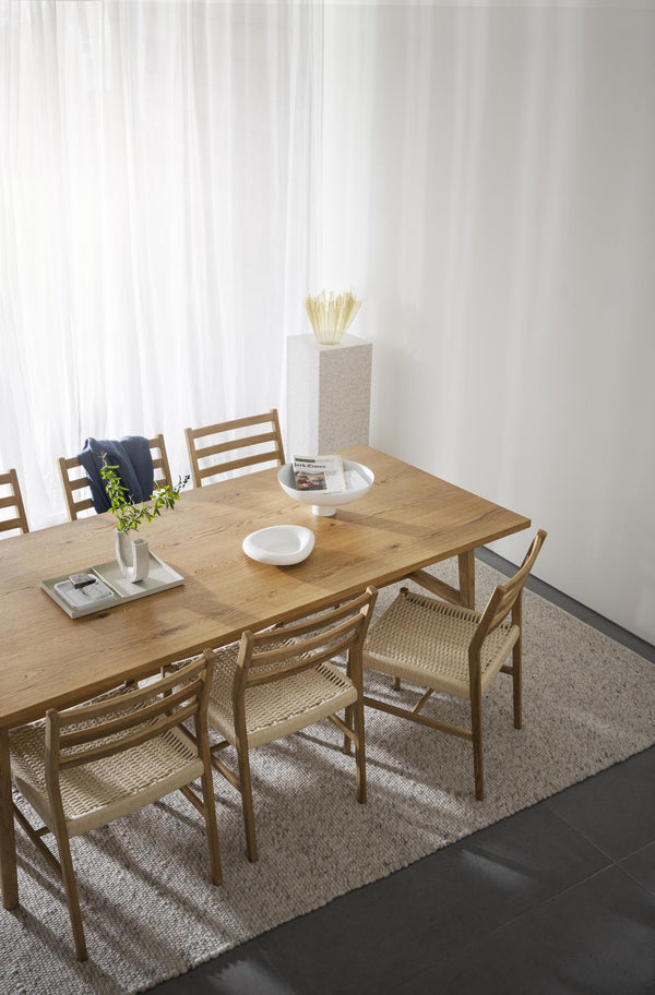 Matbord i ek med sex stolar i rotting, vit pedestal med torkade blommor och ljus gardin i bakgrunden