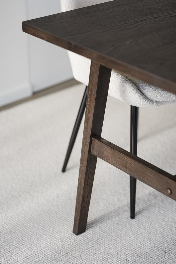 Närbild på mörkt träbord med snedställda ben och träförstärkning, stol med ljusgrått tyg i bakgrunden