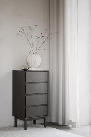 Mörk träbyrå med fyra lådor och vita dekorationer i minimalistiskt sovrum med ljusa gardiner