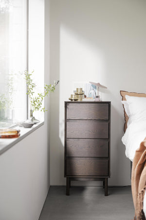 Mörkbrun sängbord med fyra lådor i trä bredvid säng med vit sängkläder och fönster med växter