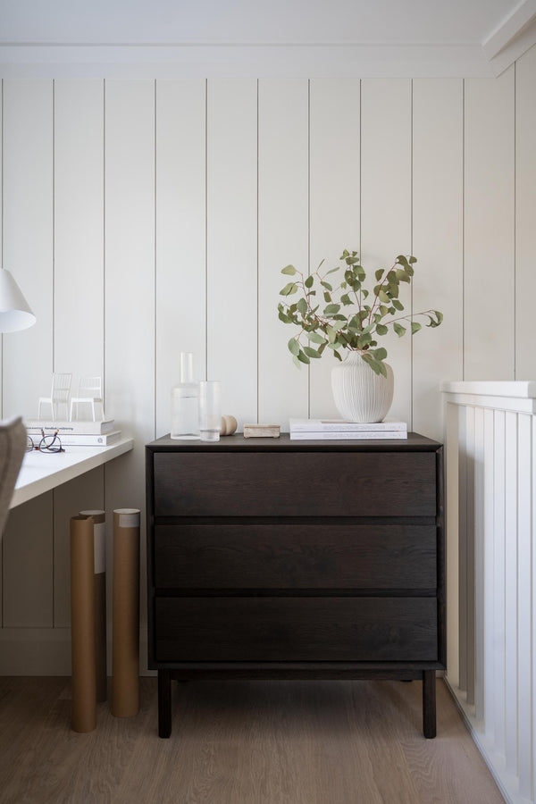 Mörk byrå med tre lådor i modern skandinavisk stil, dekorerad med vit vas och gröna kvistar