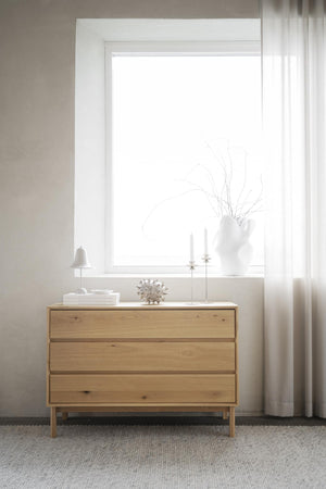 Byrå i ljust trä med tre lådor framför fönster i minimalistiskt sovrum med vita väggar och gardiner