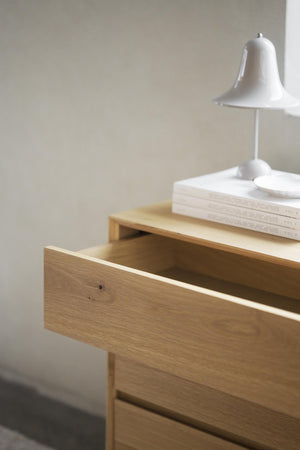 Sängbord i ljust trä med utdragen låda, vita böcker och vit lampa på toppen