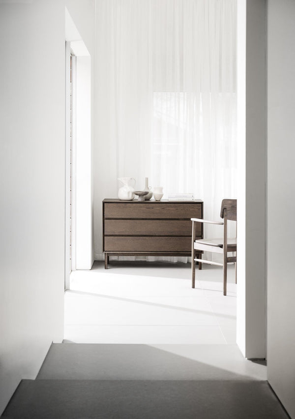 Minimalistisk inredning med mörk träkommod och stol i ljust rum med vita väggar