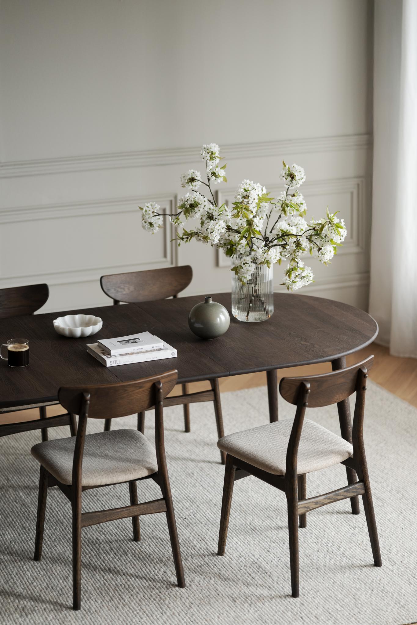 Oval matbord i mörkt trä med matchande stolar och beige klädsel, dekorerat med vita blommor i vas