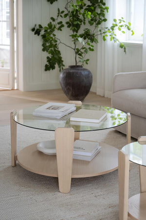 Runt soffbord i ljust trä med glasskiva och hylla, dekorerat med böcker och växt i bakgrunden
