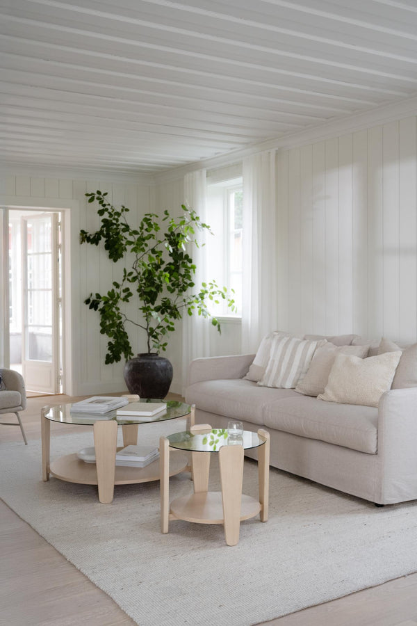 Ljust vardagsrum med beige soffa, runda soffbord i trä med glasskiva och gröna växter vid vit panel